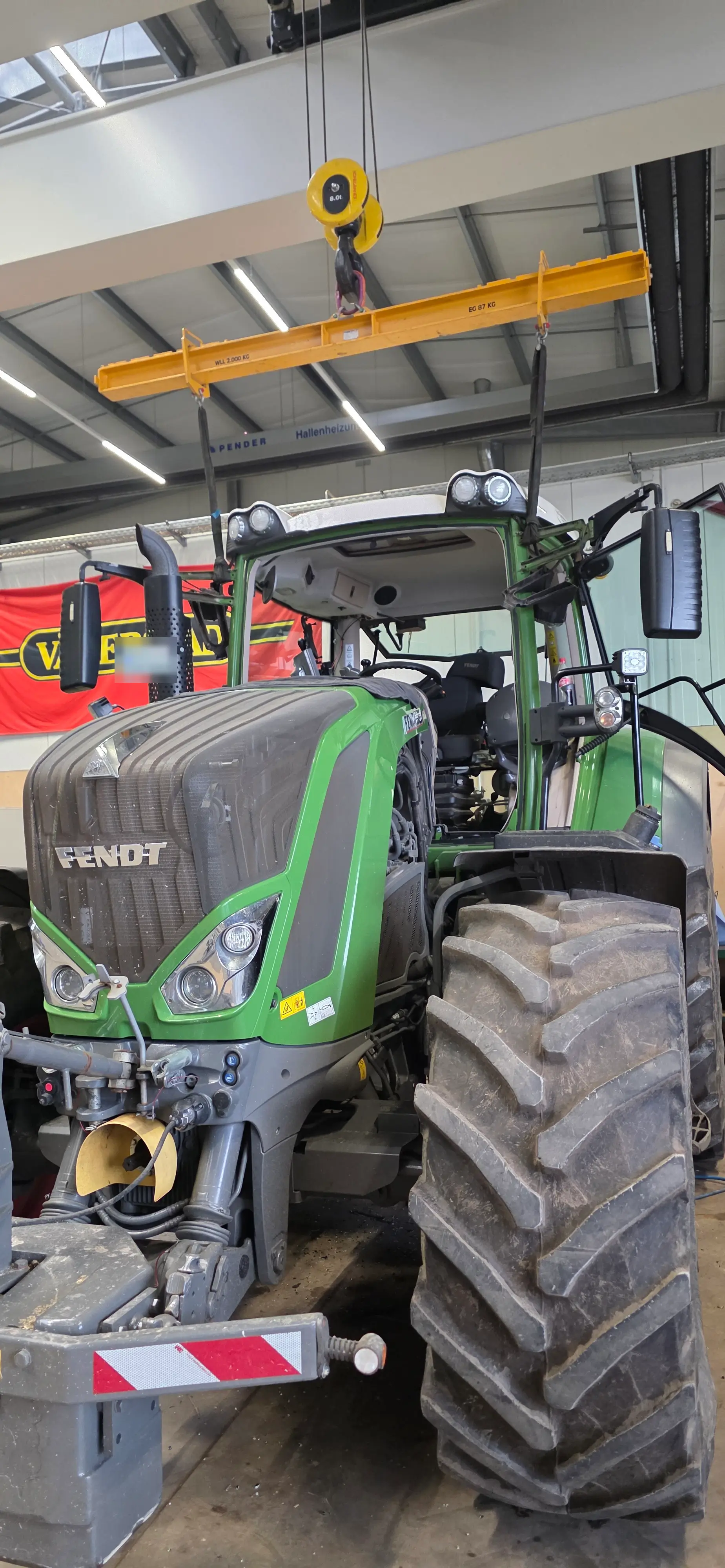 Ein Fendt Traktor ohne Frontscheibe steht in einer Werkstatt mit einem Kran über dem Fahrzeug.